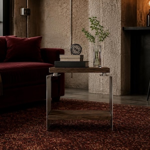 Op de afbeelding: Een bijzettafel met twee rechthoekige planken van donker hout, ondersteund door een frame van geborsteld metaal. Op de bovenste plank liggen twee boeken, een kleine zwarte geometrische draadbol en een glazen vaas met groene takken.