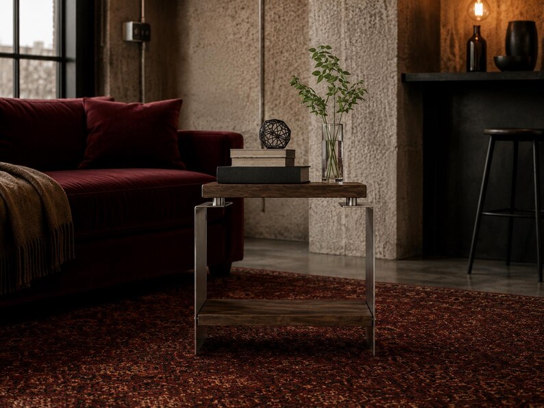Op de afbeelding: Een bijzettafel met twee rechthoekige planken van donker hout, ondersteund door een frame van geborsteld metaal. Op de bovenste plank liggen twee boeken, een kleine zwarte geometrische draadbol en een glazen vaas met groene takken.