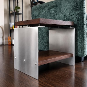 Op de afbeelding: Een bijzettafel met twee donkerbruine houten planken gescheiden door een frame van geborsteld zilverkleurig metaal. De bovenste plank wordt ondersteund door kleine metalen afstandhouders, terwijl de onderste plank met zichtbare schroeven is bevestigd.