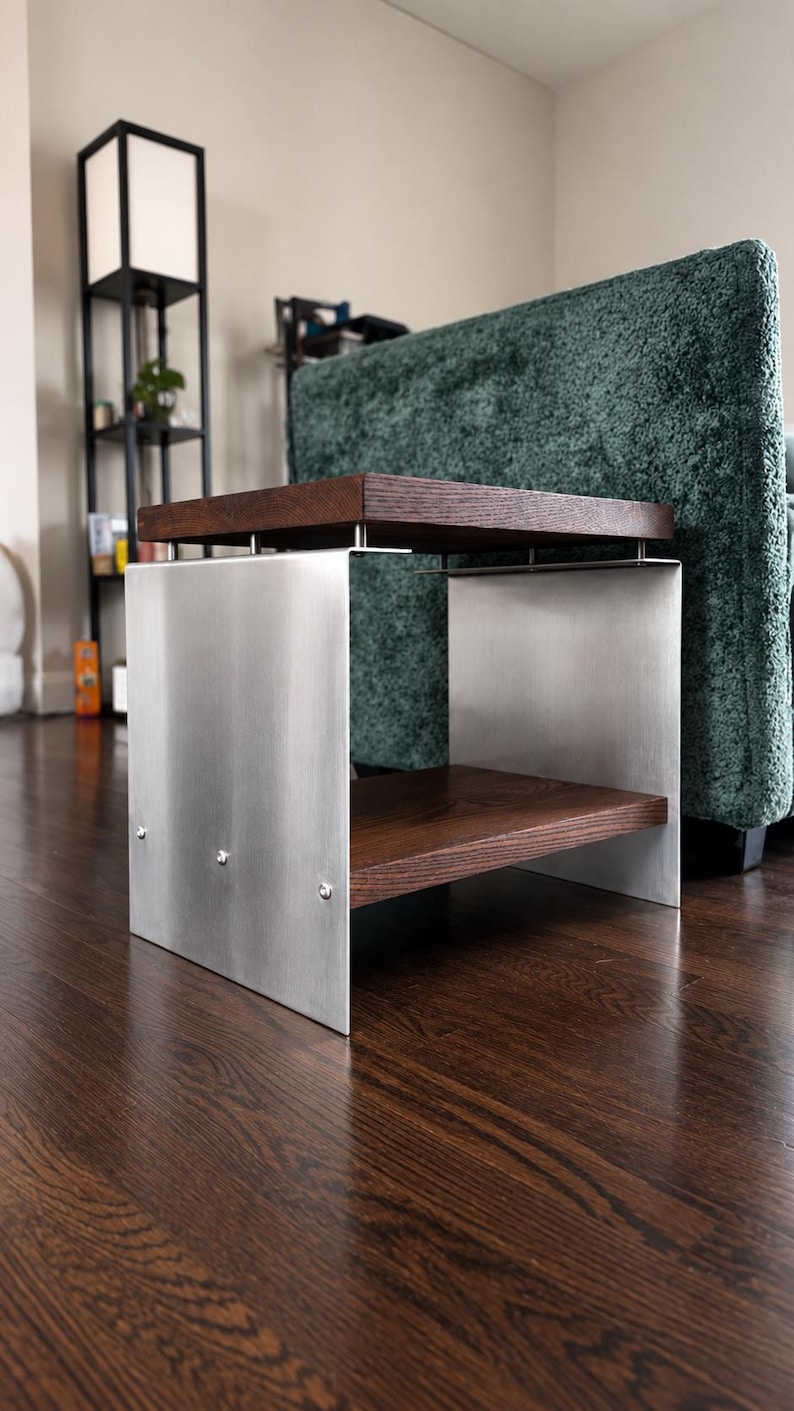 Op de afbeelding: Een bijzettafel met twee donkerbruine houten planken gescheiden door een frame van geborsteld zilverkleurig metaal. De bovenste plank wordt ondersteund door kleine metalen afstandhouders, terwijl de onderste plank met zichtbare schroeven is bevestigd.