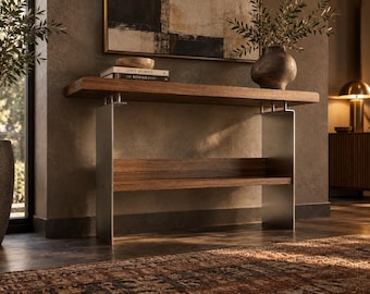 Op de afbeelding: Een consoletafel met een dik rechthoekig houten blad en een onderste houten plank, ondersteund door twee verticale poten van geborsteld metaal. De tafel heeft een strak, lineair ontwerp met zichtbare houtnerf en metalen bevestigingen.