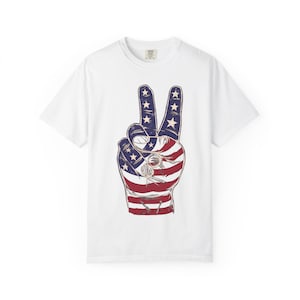 Könnte beinhalten: Ein weißes Kurzarm-T-Shirt mit einer Grafik einer Hand, die ein Friedenszeichen zeigt. Die Hand ist mit einem rot-weiß-blauen US-Flaggenmuster gefüllt, das weiße Sterne auf blauem Grund und horizontale rote und weiße Streifen aufweist.