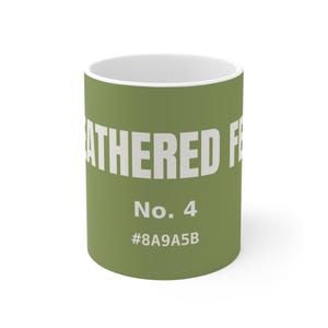 May include: A cylindrical ceramic mug with a matte olive green exterior and a white interior. The text FEATHERED FE... is printed in large white capital letters, followed by the text No. 4 and the color hex code #8A9A5B in white font.