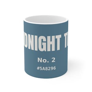 Op de afbeelding: Een cilindervormige keramische mok met een matte blauwe buitenkant en een witte binnenkant. De tekst MIDNIGHT T staat in grote witte hoofdletters, gevolgd door de tekst No. 2 en de hex-kleurcode #5A8296 in het wit op de voorzijde.