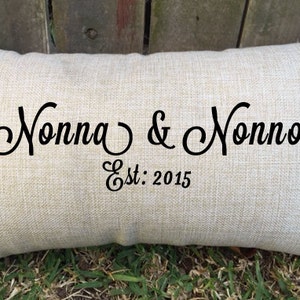 May include: A beige linen pillow with black cursive text that reads "Nonna & Nonno Est: 2015".