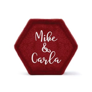 May include: A burgundy velvet hexagonal ring box with the names "Mike & Carla" in white script. The box is a deep red color and has a soft, textured appearance. The design is simple and elegant.
