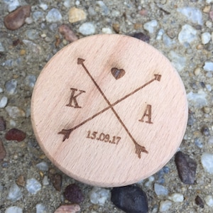 Puede incluir: Un disco redondo de madera con un corazón y dos flechas grabados en él. Las letras K y A también están grabadas en el disco, junto con la fecha 15.09.17.