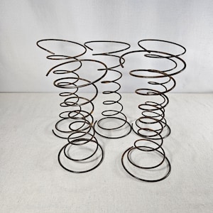May include: Five rusty metal springs, likely from a bed frame, are arranged in a group on a white surface. The springs are coiled and have a slightly aged appearance.