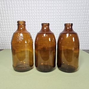 May include: Three brown glass bottles with rounded bodies and narrow necks. The bottles are empty and have a vintage appearance. One bottle has a decorative embossed design. The bottles are arranged in a row.