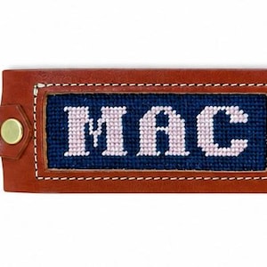 May include: A brown leather keychain featuring a rectangular needlepoint insert with a dark blue background and the light pink text MAC stitched in the center. The leather is secured with two brass-colored metal rivets and a silver-toned metal ring.