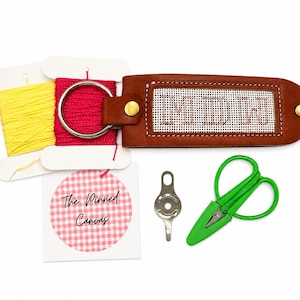 May include: A brown leather keychain with a needlepoint insert featuring the letters MDW, attached to a silver ring. Includes yellow and pink embroidery floss on cards, a small metal tool, green scissors, and a tag labeled The Pinned Canvas.