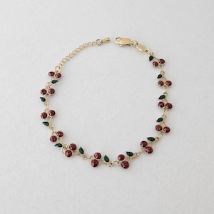 Peut inclure: Un bracelet à chaîne dorée orné d'une série de petites breloques cerises rouge foncé avec des feuilles vertes. Le bracelet comprend une fine chaîne à maillons, un fermoir mousqueton et une chaînette d'extension réglable à une extrémité.