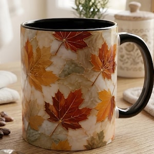 May include: A ceramic mug featuring a repeating pattern of autumn maple leaves in shades of orange, red, and yellow against a marbled white background. The mug has a solid black interior and a matching black handle, providing a high-contrast look.