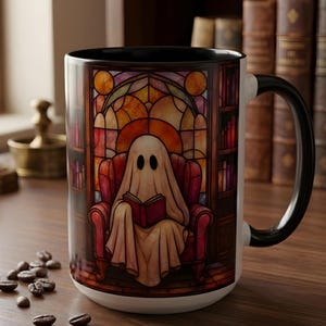 May include: A ceramic mug with a black handle and interior, featuring a printed design of a ghost draped in a white sheet sitting in a red armchair reading a red book, set against a background of a library with stained glass window panels.