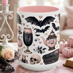 May include: A white ceramic mug with a pink interior featuring a repeating pattern of black bats, pink pumpkins, a skull, a coffin, a teapot, a wine glass, and small pink hearts scattered across the exterior surface of the cylindrical vessel.
