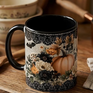 May include: A ceramic mug with a black handle and interior, featuring a decorative black lace border at the top and bottom. The exterior displays a printed design of a large orange pumpkin, assorted white and dark flowers, and a small black bat.