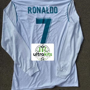 May include: A white long-sleeved football shirt featuring the name RONALDO and the number 7 in teal lettering on the back. A rectangular white logo with the text ultrakits SOCCER JERSEYS ONLINE and a football graphic is positioned centrally on the shirt.