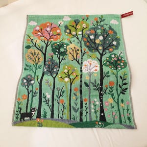 Può includere: Un asciugamano rettangolare verde menta con una scena boschiva tessuta che raffigura vari alberi colorati, piccole nuvole bianche e un gatto bianco e nero in piedi su un sentiero. Una piccola etichetta rossa nell'angolo in alto a destra riporta il testo Shmily towel.