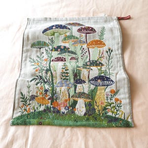 Peut inclure: Un torchon rectangulaire gris clair orné d'une broderie détaillée représentant divers champignons dans des tons de bleu, orange, violet et vert, entourés de petites fleurs sauvages et de feuillage sur une base d'herbe verte.