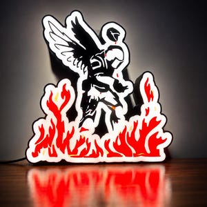 May include: A custom LED neon sign featuring a black silhouette of a winged figure in a helmet standing amidst bright red and orange flames. The light fixture has a white border and emits a vibrant glow, creating a sharp contrast against the dark.