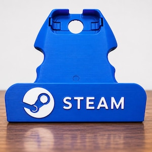 Könnte beinhalten: Ein blauer Kunststoffständer für ein tragbares Spielgerät mit dem weißen Steam-Logo und dem Wort STEAM auf der Vorderseite. Der Ständer hat eine konturierte Form mit einer zentralen kreisförmigen Aussparung und einem vertieften oberen Bereich.