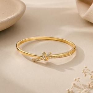May include: A gold-toned hinged bangle bracelet featuring a central knot design. The knot and one side of the band are embellished with a row of small, clear, sparkling stones. The bracelet has a polished metallic finish and a secure clasp mechanism.