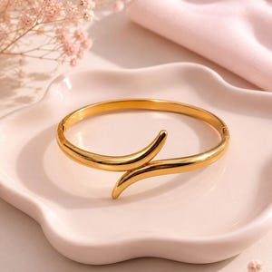 May include: A polished gold-tone metal bangle bracelet featuring a bypass design with two tapered, overlapping ends. The bracelet has a smooth, reflective surface and a hinged closure mechanism visible on the side, creating a continuous circular form.