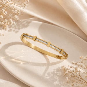 May include: A gold-toned bangle bracelet featuring a central channel set with a row of small, clear, rectangular-cut stones. The metal band is accented by three evenly spaced vertical gold bars that divide the row of stones into four segments.