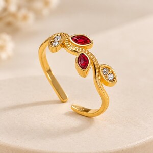 May include: A gold-toned open ring featuring a vine-like design with two marquise-cut red stones and two clusters of small, clear round stones. The band has a textured, beaded surface detail that mimics the appearance of a delicate plant stem.