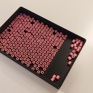 May include: A rectangular black sorting tray filled with numerous small, cylindrical pink beads. The beads are arranged in a grid pattern within the main section of the tray, while a few loose beads are scattered in the open space on the right side.
