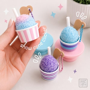 May include: Three snow cone clickers featuring textured, colourful shaved ice tops in pink and blue, solid blue, and purple. Each includes a white straw, a tan heart-shaped charm, and a small ball chain. The cups have striped patterns.