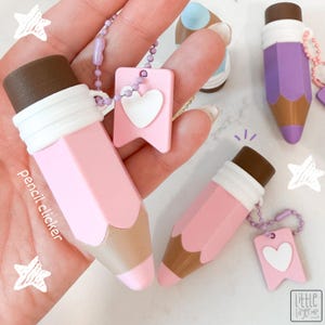 Peut inclure: Un clicker en forme de crayon rose et marron avec une virole blanche et une chaînette à billes violet clair munie d'une étiquette rectangulaire rose avec un cœur blanc. Le texte "pencil clicker" est visible à gauche et le logo "little layer" en bas.