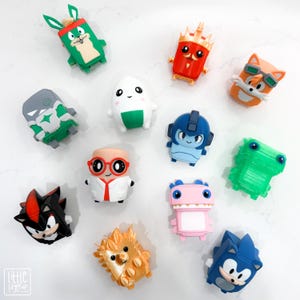 May include: A collection of twelve small, stylised plastic character figurines featuring various colours and designs, including a white egg-shaped figure, a blue robot, a black and red hedgehog, a pink dinosaur, and an orange fox with goggles.
