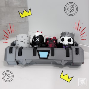 May include: A grey rectangular container holding four small stylised figurines. The figures include a white pillow-shaped character with stars, a black character with a white mohawk, a red and black patterned figure, and a skull-faced figure with a crown.