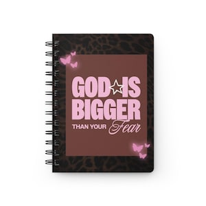 Puede incluir: Un cuaderno de espiral con una cubierta de estampado de leopardo marrón oscuro. Un panel central rectangular marrón presenta el texto rosa "GOD IS BIGGER THAN YOUR FEAR" con un icono de estrella y varias mariposas rosas brillantes alrededor.