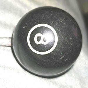 May include: A spherical black gear shift knob featuring a white number eight inside a white circle on the top surface. The knob has a metallic threaded bolt protruding from the side, designed for attachment to a gear stick or similar mechanism.