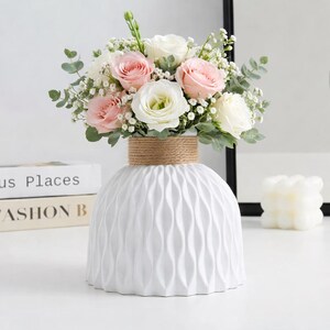 May include: A white vase with a vertical wavy texture and a natural jute rope wrapped around the neck. The vase contains a floral arrangement of light pink roses, white roses, small white baby's breath flowers, and green eucalyptus leaves.