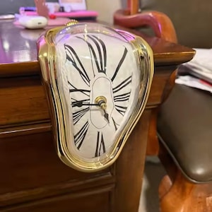 May include: A melting-style analog clock with a gold-toned frame and a white face featuring black Roman numerals. The clock is designed to drape over the edge of a flat surface, with black hour and minute hands centered on the dial.