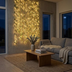 May include: A wall-mounted string light curtain featuring artificial green ivy leaves intertwined with warm white LED lights. The strands hang vertically against a concrete wall, creating a glowing, illuminated backdrop of foliage and light.