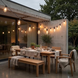 Puede incluir: Un conjunto de comedor de exterior de madera con una mesa rectangular, un banco a juego y sillas con mantas de colores claros. Una guirnalda de bombillas estilo Edison cuelga sobre la mesa, iluminando el patio con un brillo cálido.