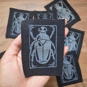 May include: A rectangular black fabric patch featuring a screen-printed light grey illustration of a beetle centred within a rectangular border. The patch has raw, unfinished edges and is shown in a collection of identical pieces.