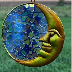 May include: A gold crescent moon wind chime with a mosaic of blue, green, and gold glass pieces. The moon has a smiling face.