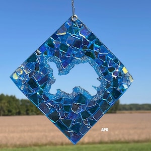 May include: A diamond-shaped suncatcher with a mosaic design in shades of blue and green. The center features a cutout shape, and the piece is suspended by a silver chain. The letters "APD" are visible in the lower right corner.
