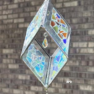May include: A geometric, stained glass hanging decoration. The clear glass structure is diamond-shaped with colorful mosaic glass accents in blue, orange, and green. Two iridescent teardrop-shaped crystals dangle from the bottom and center.