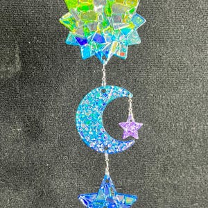 May include: A celestial-themed hanging decoration featuring a starburst, crescent moon, and star. The starburst at the top has green, yellow, and blue hues. The moon and star are blue with glitter, and the star is purple. All pieces are connected by silver chains.