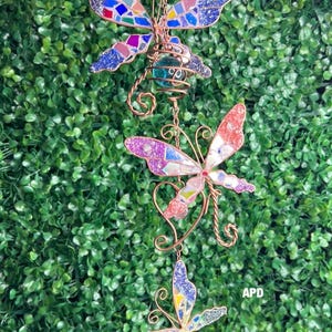 May include: A decorative hanging butterfly mobile with three butterflies. Each butterfly has colorful mosaic-style wings in shades of blue, purple, and red. The butterflies are connected by copper wire and hang against a green background.