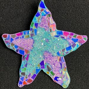 May include: A mosaic starfish ornament with a shimmering, iridescent design. The ornament features a central area of light blue glitter, surrounded by purple glitter and colorful mosaic pieces. The edges are outlined with a silver wire for hanging.