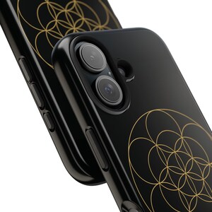 May include: Two black smartphone cases featuring a gold geometric pattern of overlapping circles on the back. The cases have cutouts for a dual-lens camera system and side buttons. The design is printed on a smooth, dark, solid-colored surface.