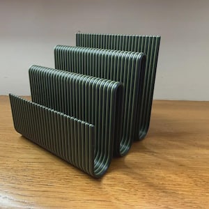 May include: A dark green letter holder featuring a tiered, U-shaped design with three distinct compartments. The structure is composed of multiple parallel vertical ridges that create a textured, ribbed appearance across the entire surface.