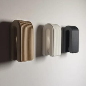 May include: Three wall-mounted hooks in a U-shape design with vertical ribbed textures. The hooks are arranged in a row, featuring a tan hook on the left, a white hook in the center, and a black hook on the right, all attached to a flat surface.